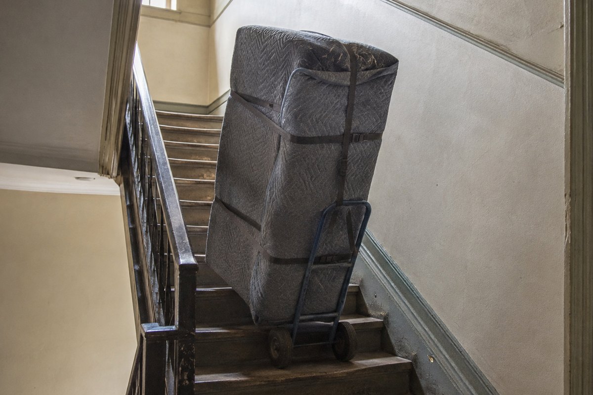 Ein in graue Decken gewickelter Zweisitzer steht aufrecht festgegurtet auf einer Sackkarre im Treppenhaus eines Altbaus.